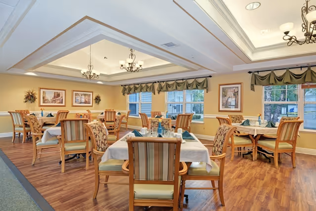 Bright, elegant dining room with multiple set tables and wooden chairs beneath decorative chandeliers.