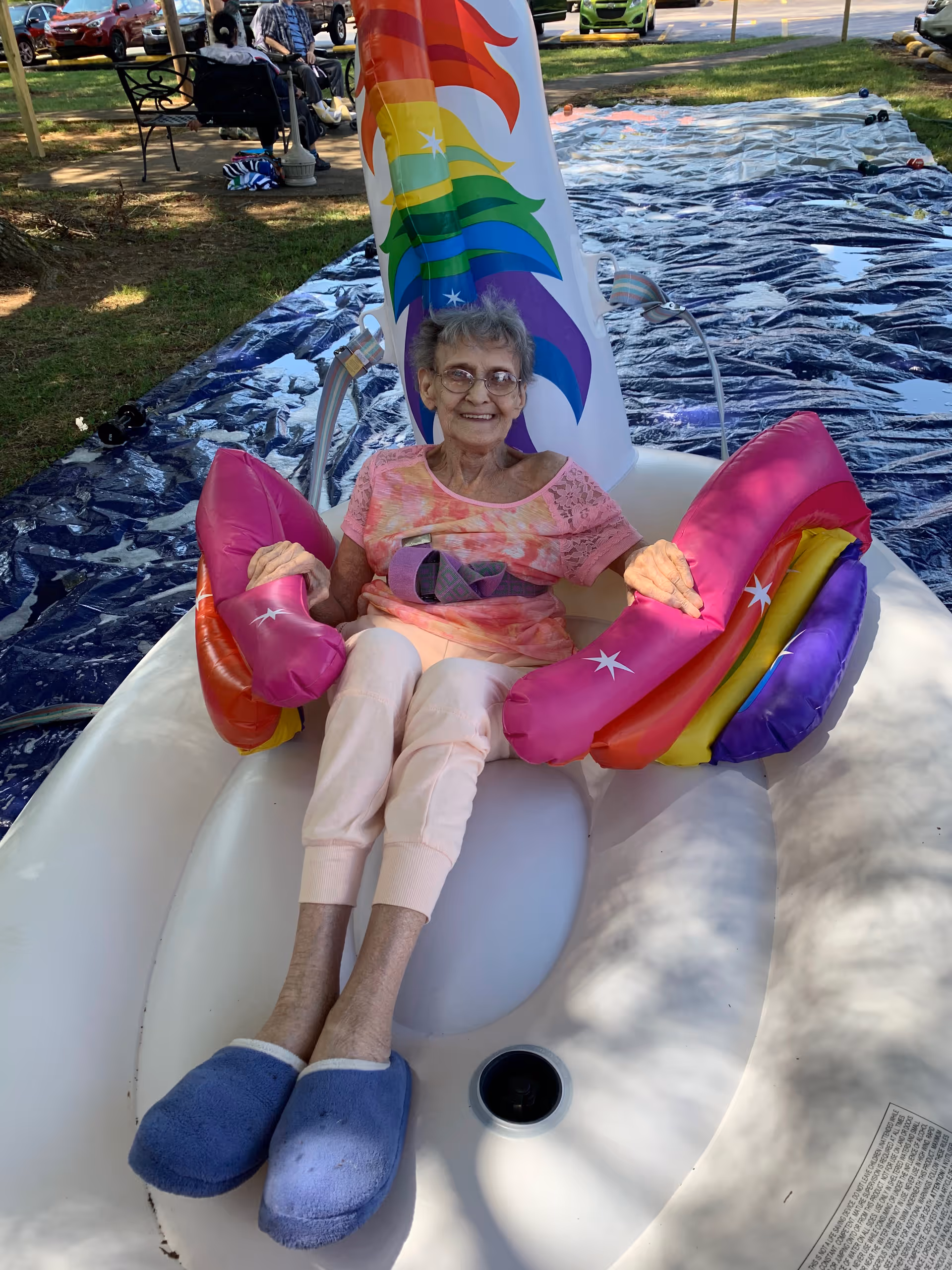 An elderly woman wearing glasses, a pink patterned shirt, light pink pants, and blue slippers is sitting and smiling on a white inflatable float with rainbow-colored armrests and a rainbow sail. The float is placed on a blue tarp outdoors with grass and trees visible in the background. There are people sitting on benches and parked cars in the background.