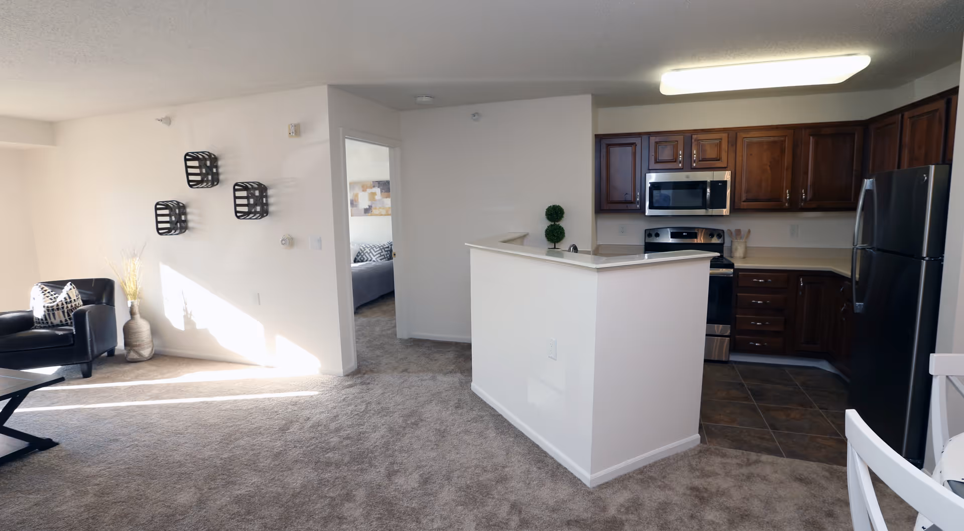 Interior view of a retirement community apartment showing a small kitchen with dark wooden cabinets, stainless steel appliances including a refrigerator, stove, and microwave. The kitchen has tiled flooring and a white half-wall separating it from the carpeted living area. The living area features a black armchair with a patterned pillow, a coffee table, and decorative wall shelves. A doorway leads to a bedroom with a bed visible inside.