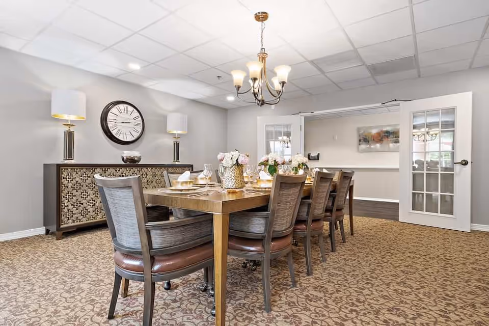A formal dining room with a long wooden table set for a meal, upholstered chairs, a chandelier, and a sideboard with lamps and a wall clock.