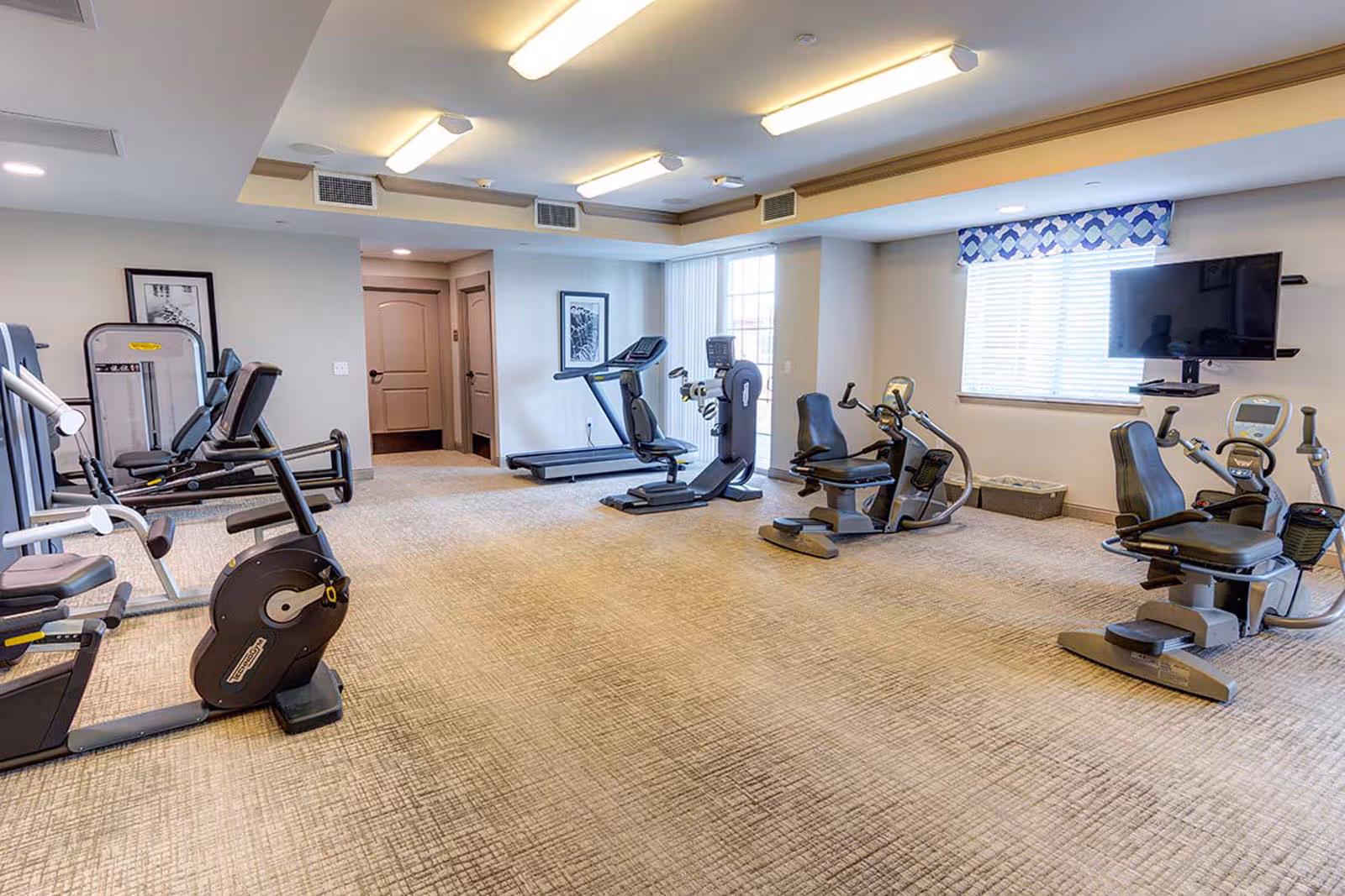 A fitness room in a senior living facility with various exercise equipment including stationary bikes, treadmills, and elliptical machines. The room has beige carpet, white walls, ceiling lights, a window with blinds and a blue patterned valance, and a wall-mounted TV.