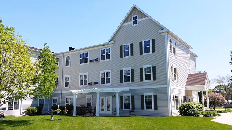 Three-story beige siding apartment building with green shutters, a small covered porch and a well-kept lawn under a clear blue sky.