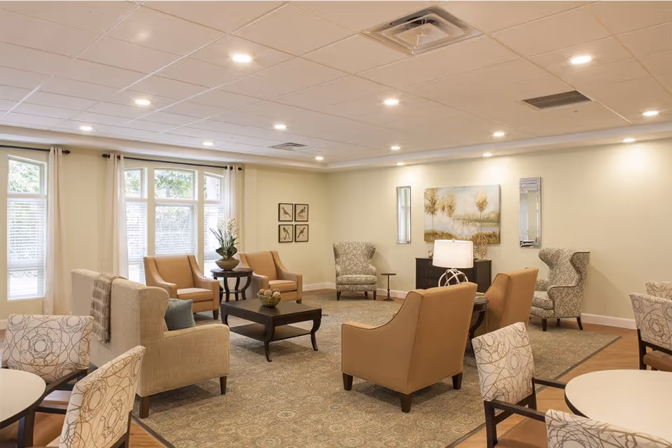 Bright common room with upholstered armchairs, side tables, and large windows.