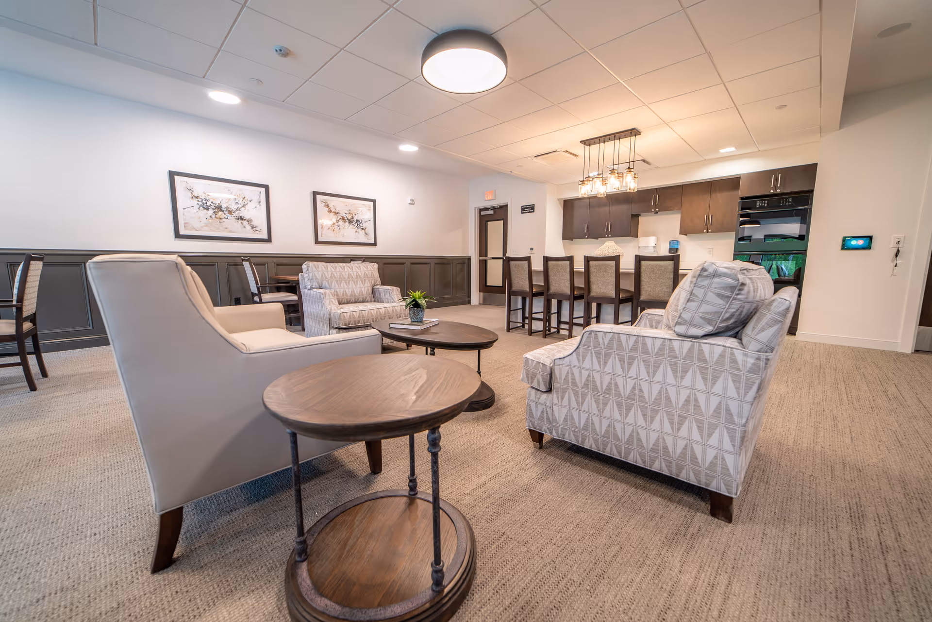 Spacious communal lounge with upholstered chairs, round wooden side tables, a coffee table, and a kitchen island with bar stools in the background.