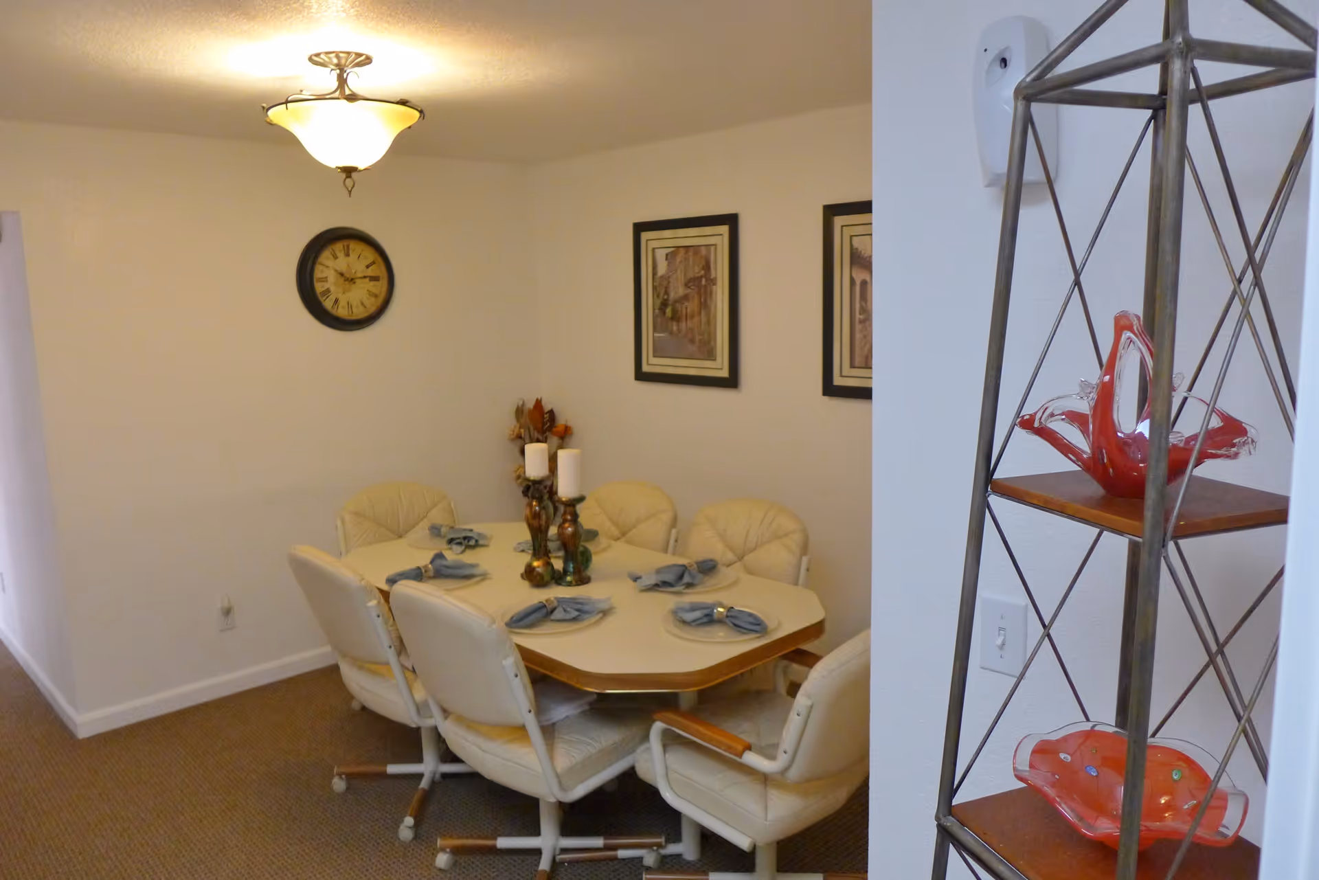 A dining room with a rectangular table set for six with beige cushioned chairs on wheels. The table has blue napkins on plates and two decorative candle holders in the center. On the wall, there is a round clock and two framed pictures. To the right, there is a metal and wood shelving unit with red decorative glass pieces.