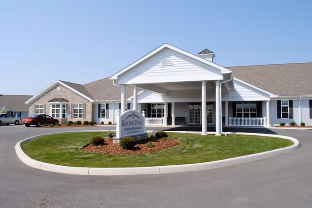 Front exterior view of Keystone Pointe Health and Rehabilitation facility with a covered entrance, manicured lawn, and a sign displaying the facility name. Several windows and a few parked cars are visible under a clear blue sky.
