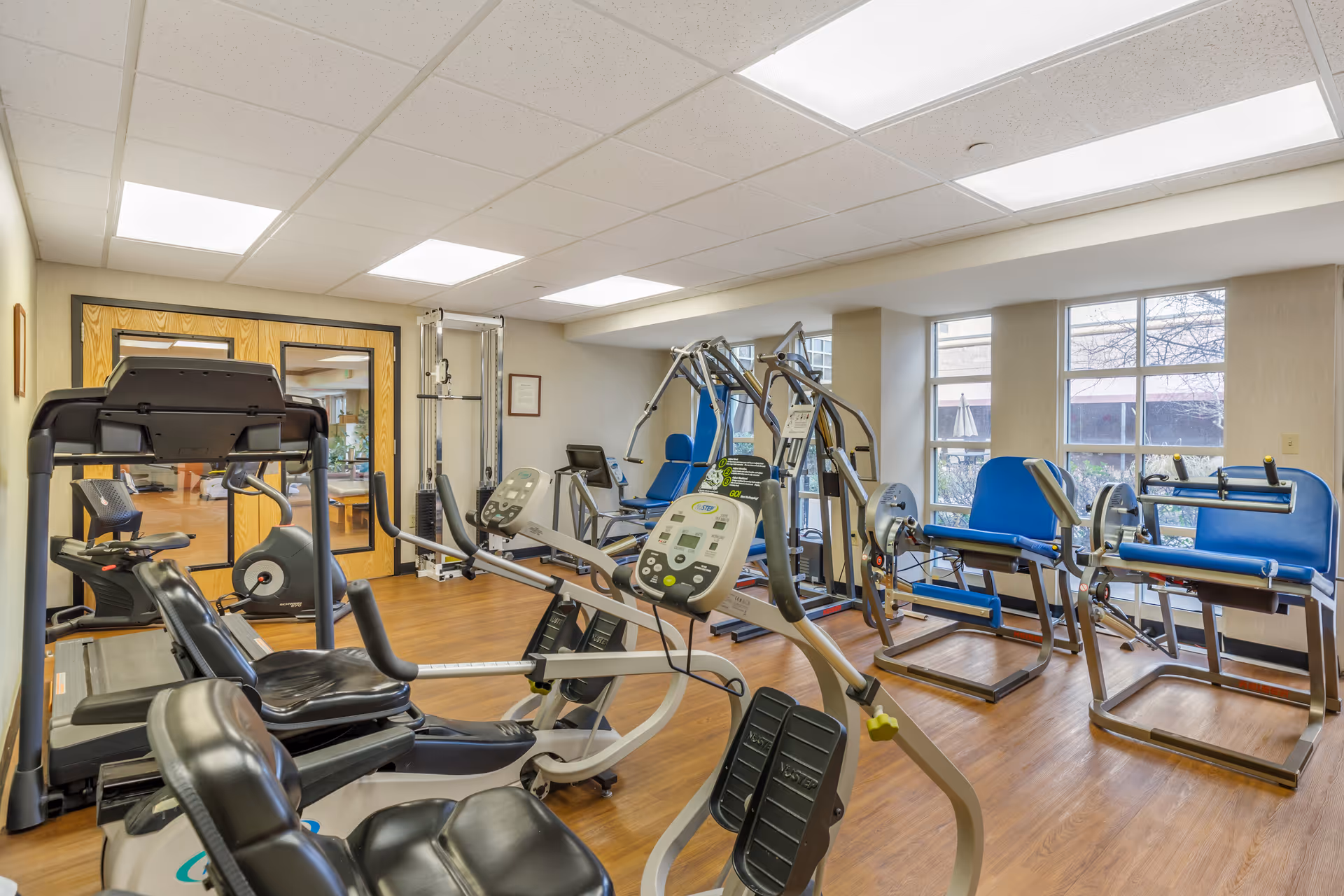 A well-lit fitness room with various exercise equipment including recumbent bikes, weight machines, and rowing machines. The room has wooden flooring, large windows letting in natural light, and a set of double wooden doors with glass panels.