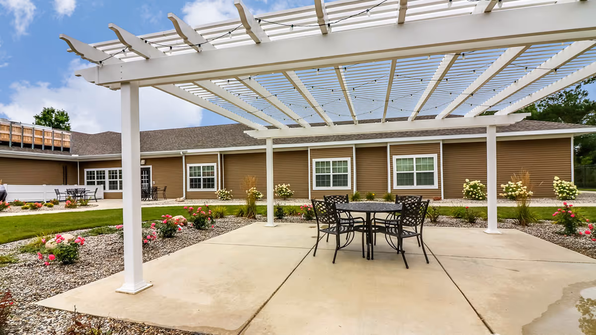 Outdoor patio area with a white pergola providing partial shade over a round metal table and four matching chairs. The patio is surrounded by landscaped flower beds with blooming pink and white flowers, and a brown building with multiple windows is visible in the background under a partly cloudy sky.