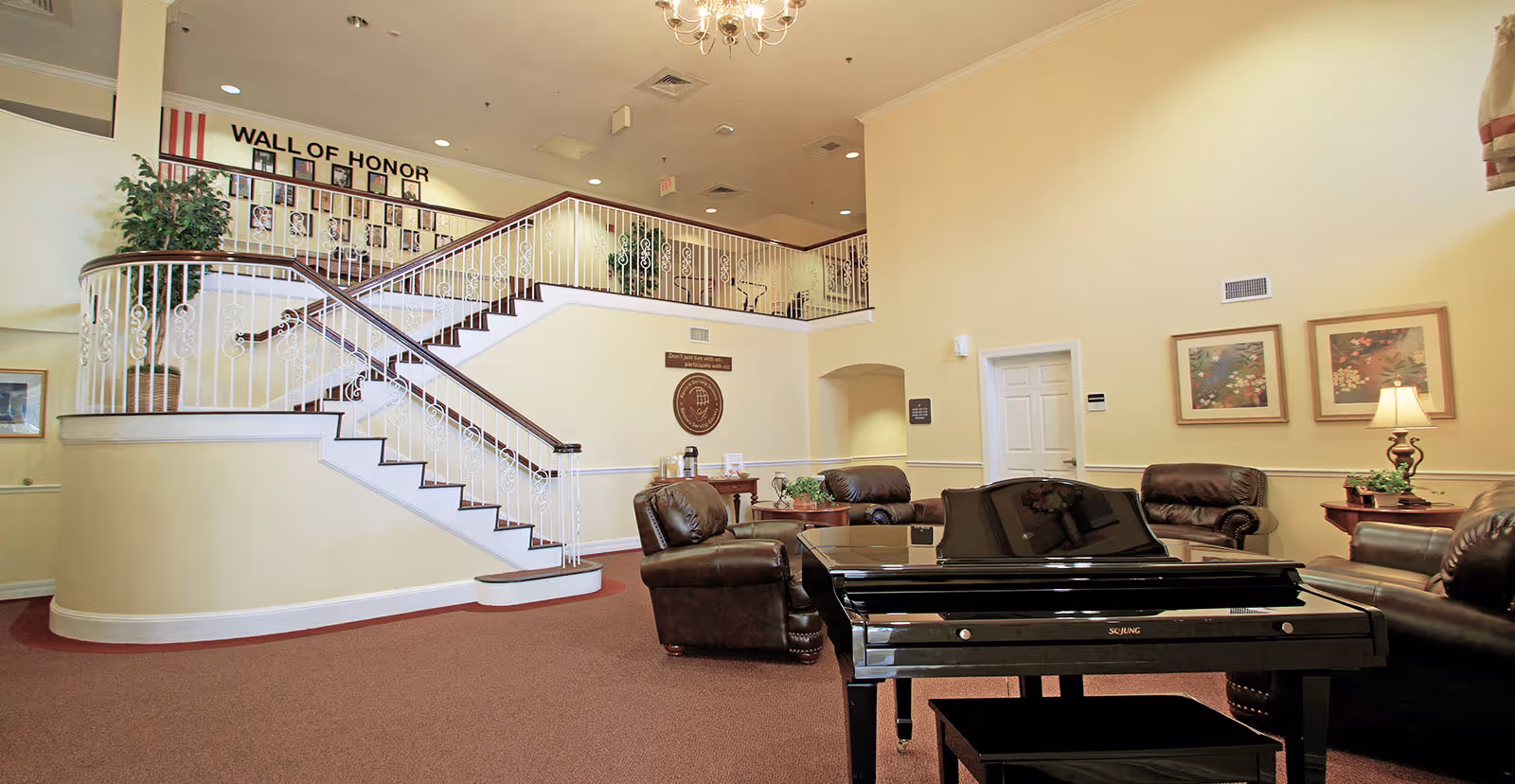 Spacious senior living common area with a grand staircase, seating cluster and a black piano under chandeliers.