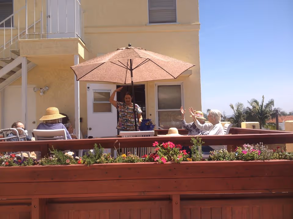 A group of elderly people sitting and socializing on a sunny outdoor patio with a large umbrella providing shade. The patio has a wooden railing with flower beds containing colorful flowers. The background shows a yellow building with windows and a door, and some palm trees are visible in the distance under a clear blue sky.