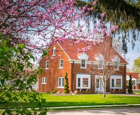 A red brick two-story building with white window frames and a red roof, surrounded by green grass and trees, some with pink blossoms, under a clear blue sky.