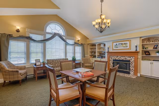 A cozy living room with a high vaulted ceiling, large arched window with blinds and drapes, a chandelier, a fireplace with a tiled surround, built-in shelves with decorative items, a wooden table with four chairs, and additional seating including armchairs and a loveseat.