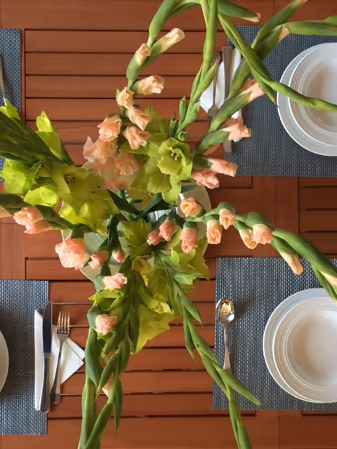 Top-down view of a wooden dining table set with blue placemats, white plates, silverware, and a centerpiece of tall green and peach flowers.