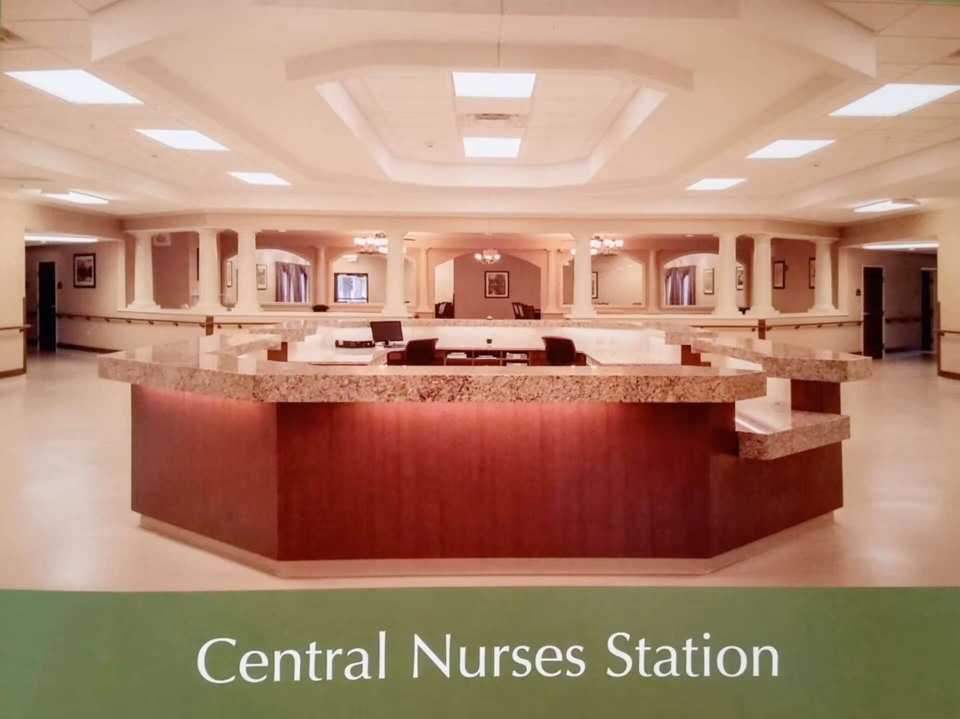 A spacious central nurses station with a large, multi-sided granite countertop and wooden base. The area is well-lit with ceiling lights and has several chairs and computer monitors inside the station. The background shows a hallway with handrails and framed pictures on the walls.
