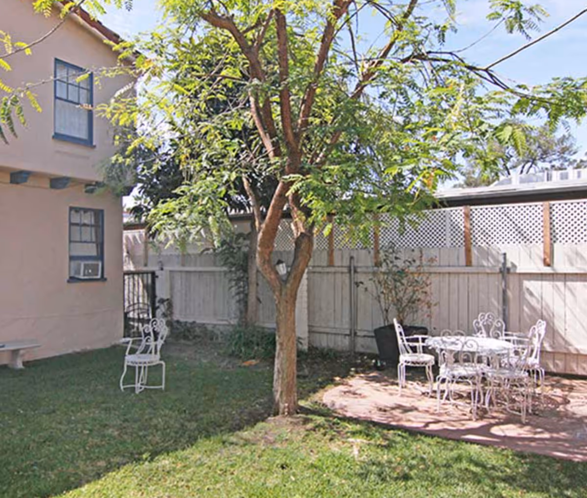 Outdoor garden area with a tree in the center, green grass, a beige building with two windows on the left, and a white metal table with four matching chairs on a paved patio area. A white wooden fence with lattice top panels encloses the space.