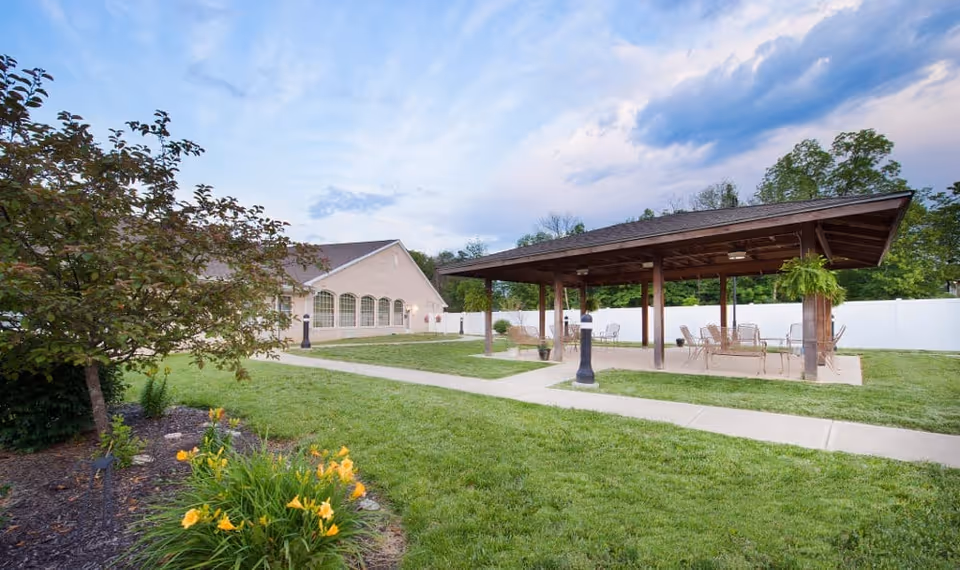 Outdoor area of Avon Health and Rehabilitation Center featuring a covered pavilion with tables and chairs, surrounded by a well-maintained lawn, flower beds, and a building with large windows in the background under a partly cloudy sky.