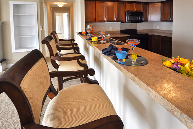 Interior view of a kitchen area with a long countertop and four wooden chairs with beige cushions lined up along the counter. The kitchen features dark wooden cabinets, a black microwave, and a stove. The countertop is decorated with colorful glass bowls and a bouquet of yellow and purple flowers. A hallway with doors is visible in the background.