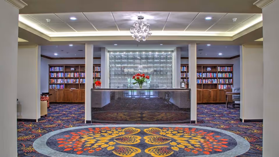 Reception desk in a senior living facility lobby with a chandelier, patterned carpet, and bookshelves behind.