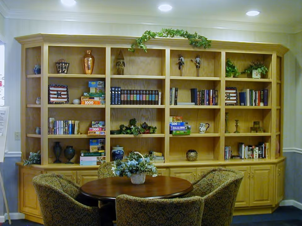 A cozy interior room featuring a large wooden bookshelf filled with books, puzzles, decorative items, and plants. In front of the bookshelf is a round wooden table with a floral centerpiece, surrounded by four upholstered chairs with patterned fabric.