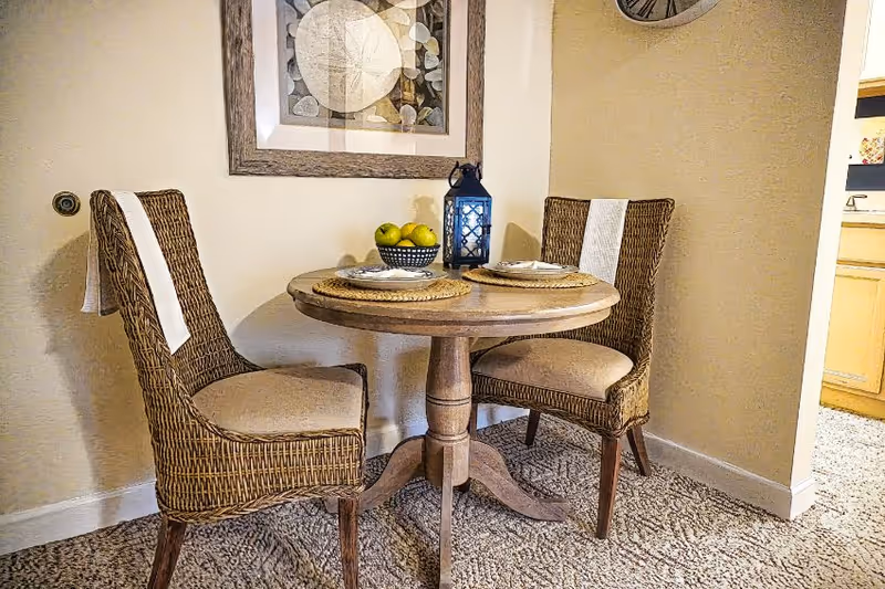 A small round wooden dining table set for two with woven chairs that have white cloths draped over the backs. On the table are two place settings, a bowl of green apples, and a decorative lantern. The room has beige walls, a framed abstract artwork, and a clock on the wall. The floor is carpeted and a kitchen area is visible through an open doorway.