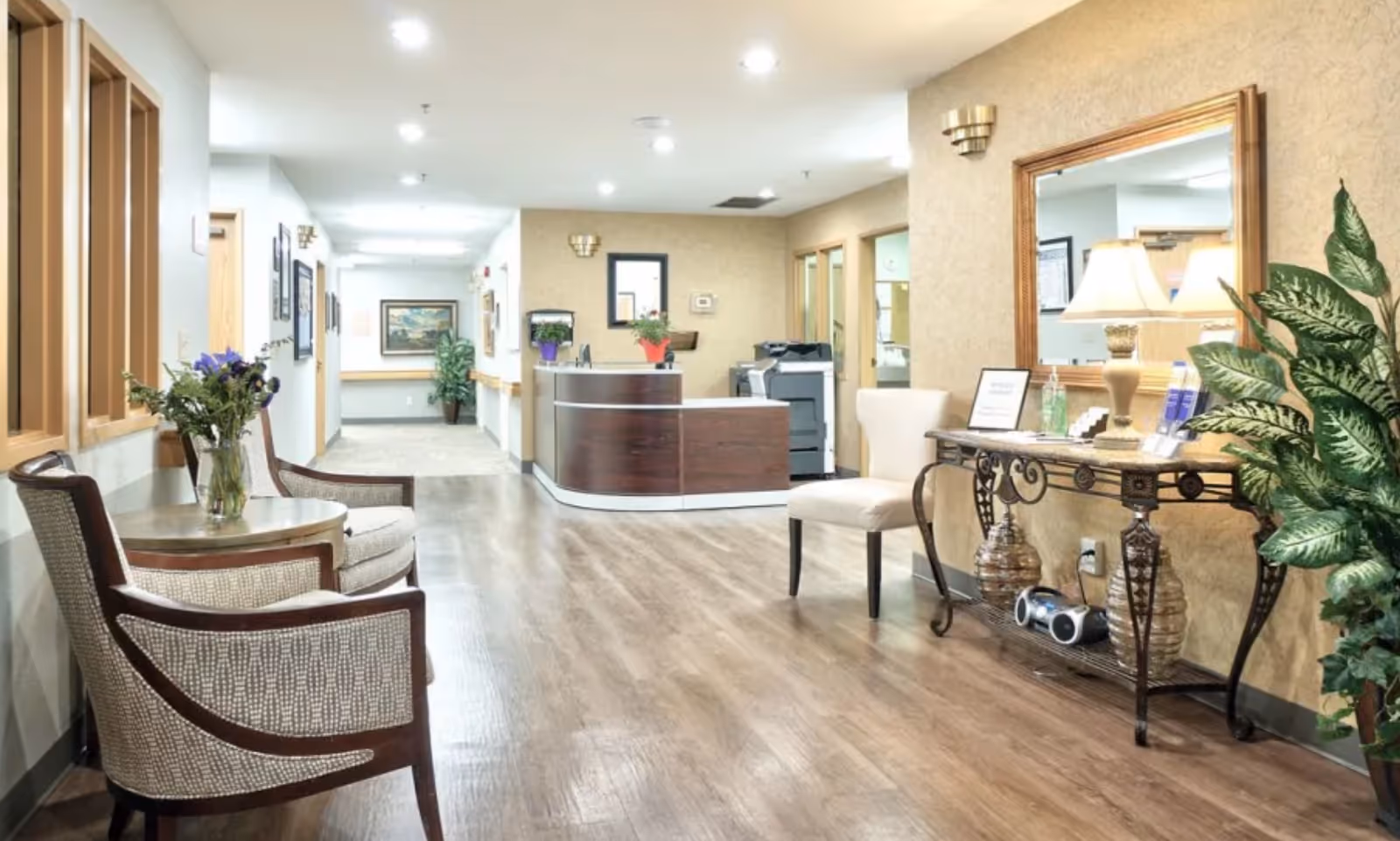 A bright and clean reception area in a senior living facility with wooden flooring, a curved wooden reception desk, two upholstered chairs with a small round table holding a vase of flowers, a decorative console table with a lamp, mirror, and plants, and a hallway leading to other rooms.