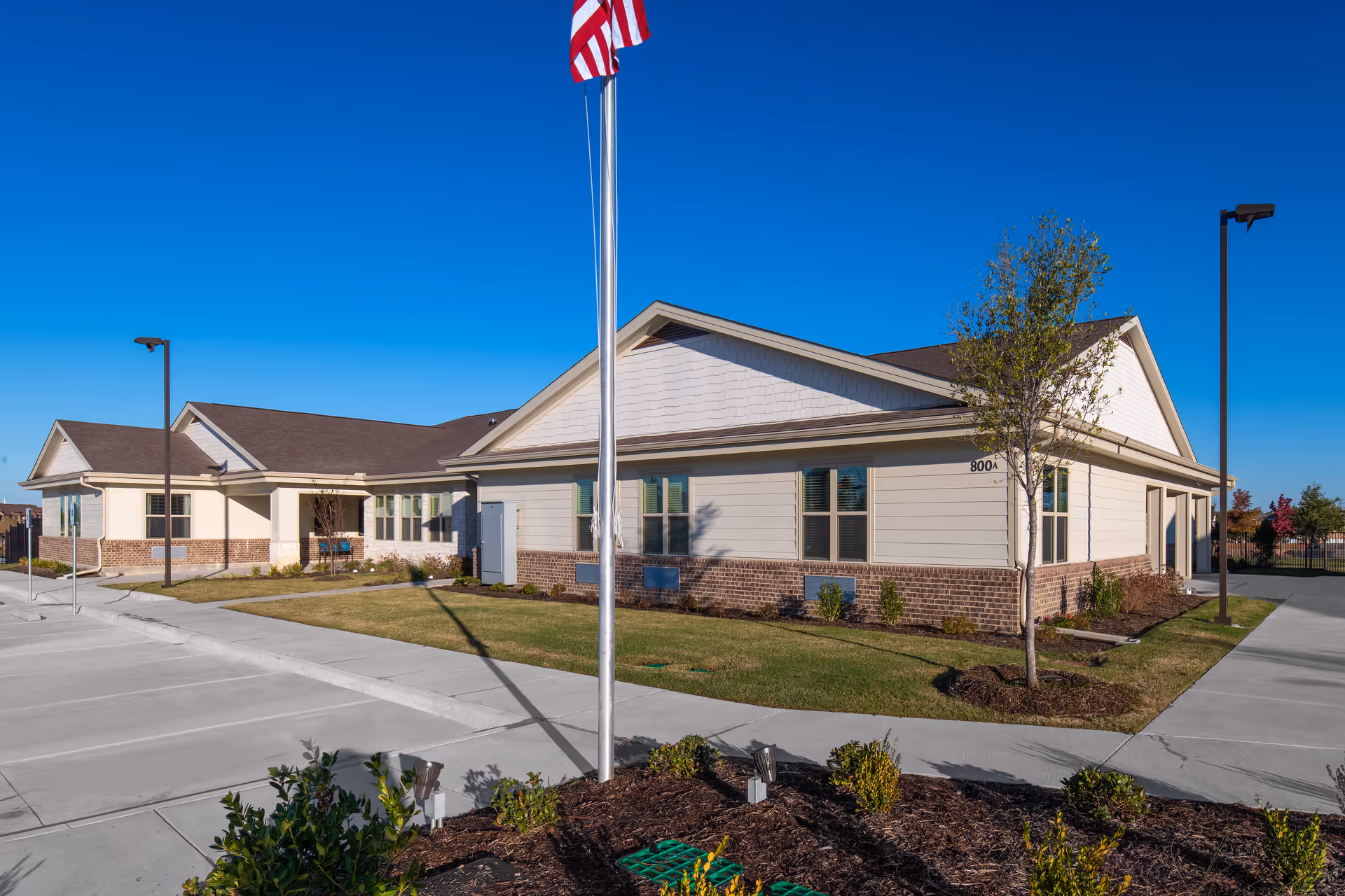 Exterior view of a single-story assisted living facility building with beige siding and brick accents, a neatly landscaped lawn, a flagpole with an American flag, and a clear blue sky.
