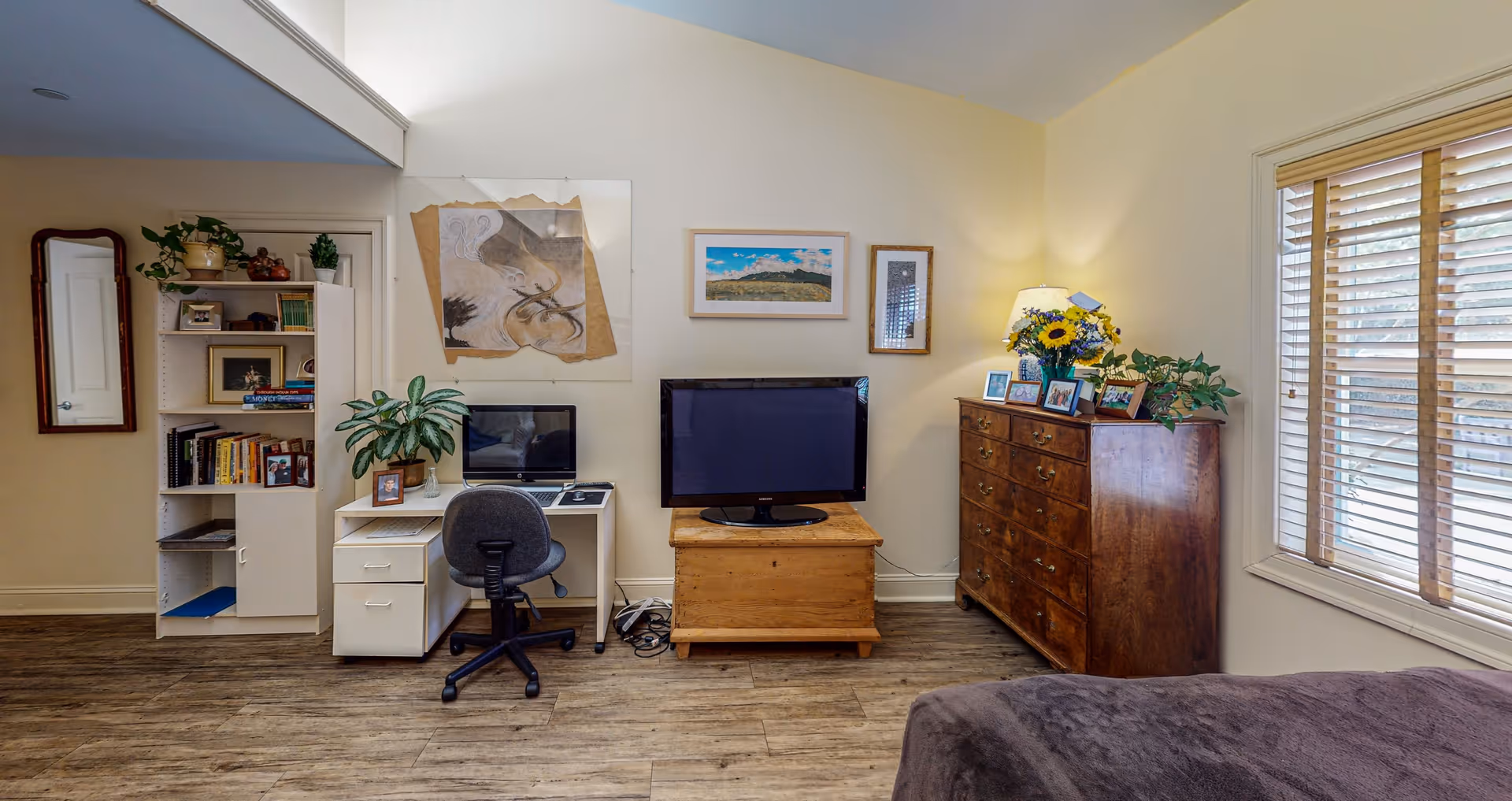 A cozy living room area with a wooden floor, a small white desk with a computer and a gray office chair, a wooden TV stand with a flat-screen TV, a wooden dresser with framed photos and a vase of flowers, a window with wooden blinds, and a bookshelf with books and decorative items.