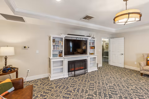 A spacious living room with a patterned carpet, a white entertainment center holding a large flat-screen TV and an electric fireplace. There are books and decorative items on the shelves. A brown leather chair with a colorful pillow is on the left, and a beige armchair is partially visible on the right. The walls are light-colored, and there is a ceiling light fixture. An open door leads to another room.
