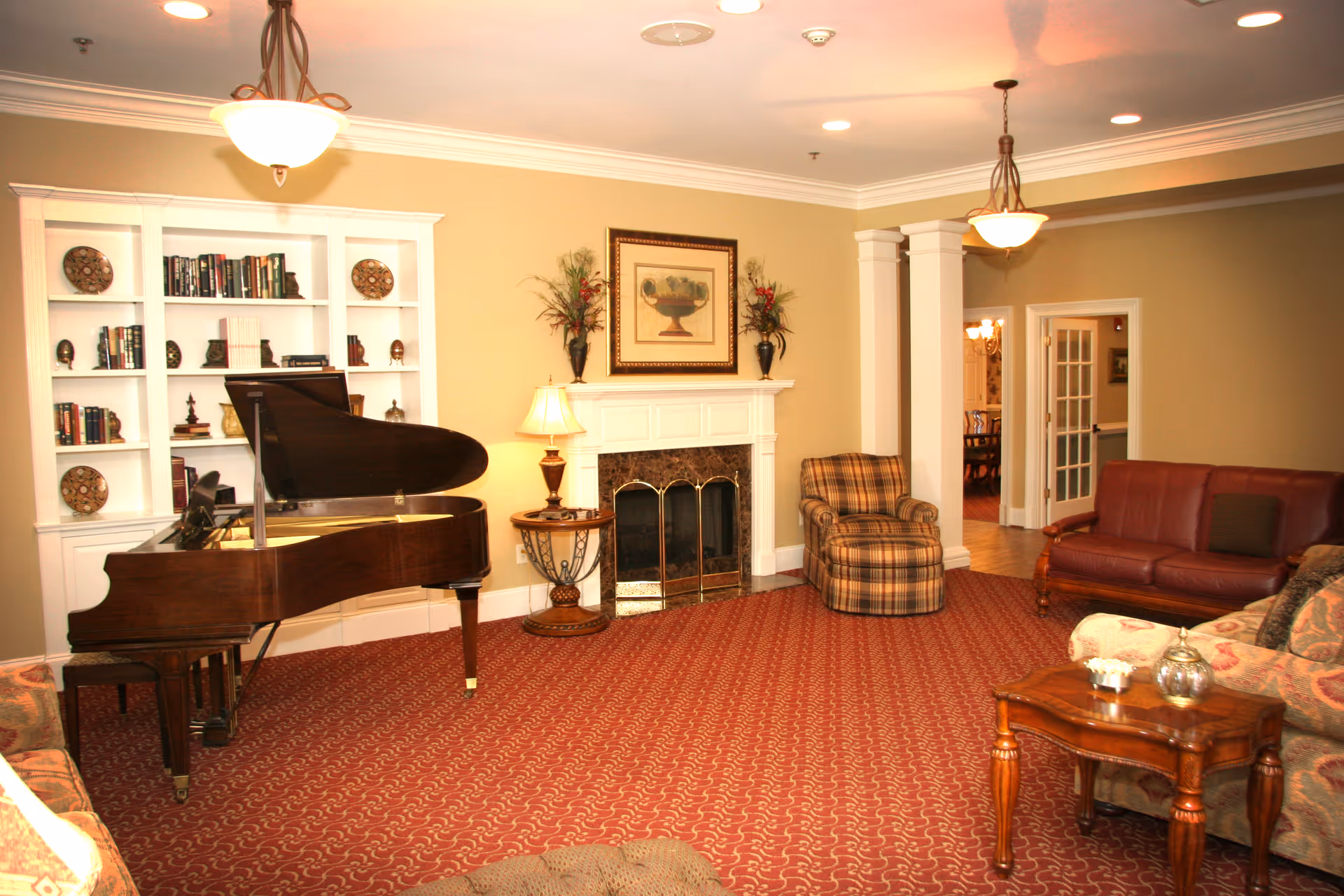 A cozy living room with a grand piano, a fireplace with a framed painting above it, a plaid armchair, a leather sofa, a floral-patterned sofa, a wooden coffee table, and built-in white bookshelves filled with books and decorative plates. The room has warm beige walls, red patterned carpet, and two ceiling light fixtures.