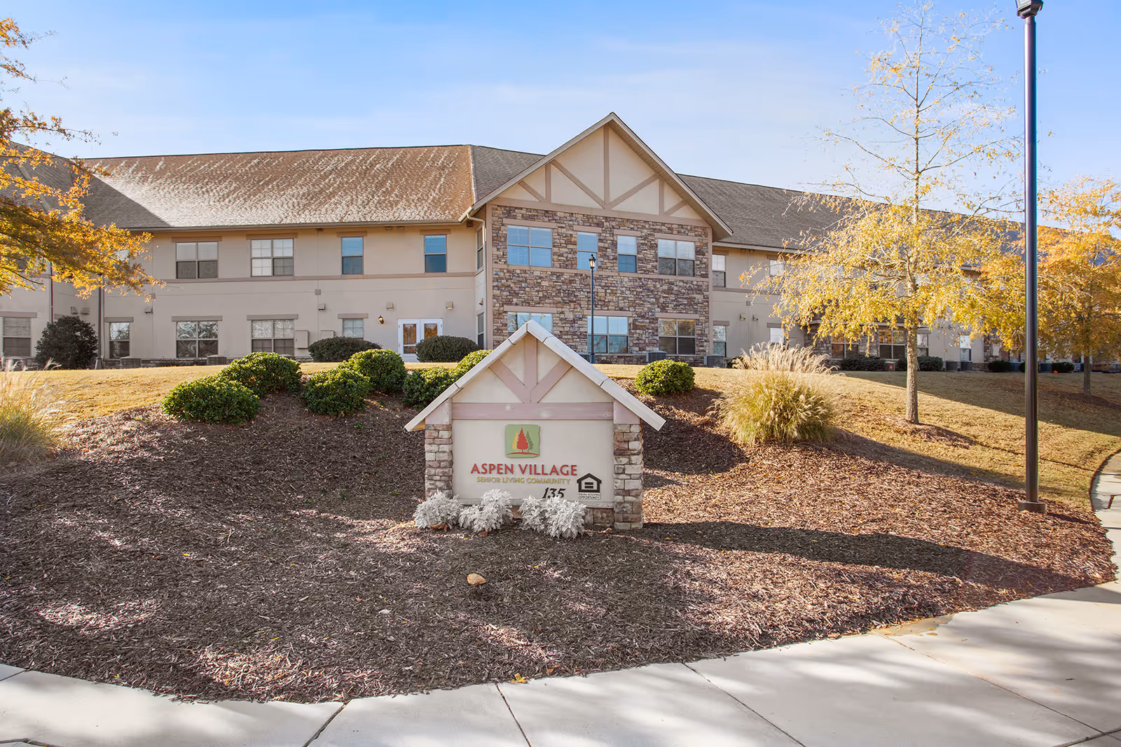 Exterior view of a senior living community building with beige and stone facade, surrounded by landscaped bushes and trees with autumn foliage. A sign in front reads 'Aspen Village Senior Living Community'.