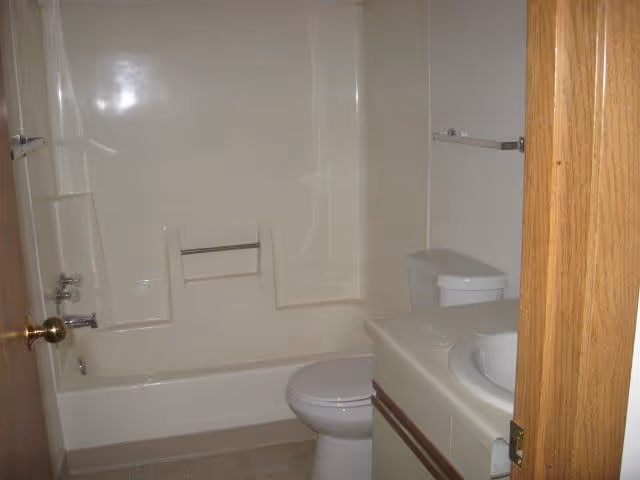 A small bathroom with a white bathtub and shower combination, a white toilet, and a white sink with a countertop. The walls and bathtub surround are light-colored, and there is a wooden door partially open showing the bathroom interior.