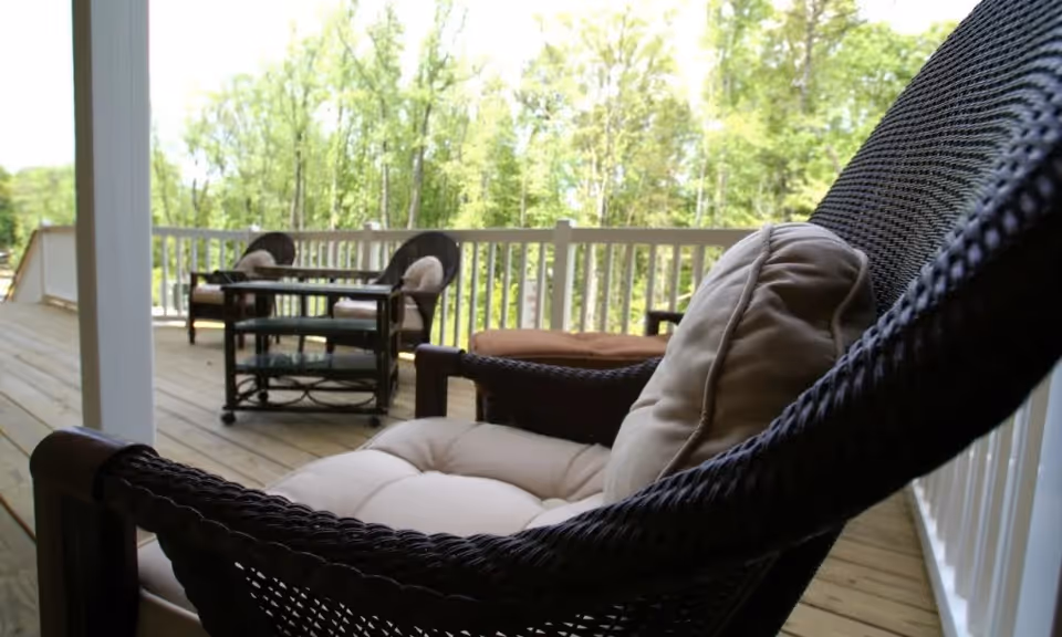 A wooden deck with cushioned wicker chairs and a small glass-top table, surrounded by a white railing and overlooking a green, tree-filled landscape.