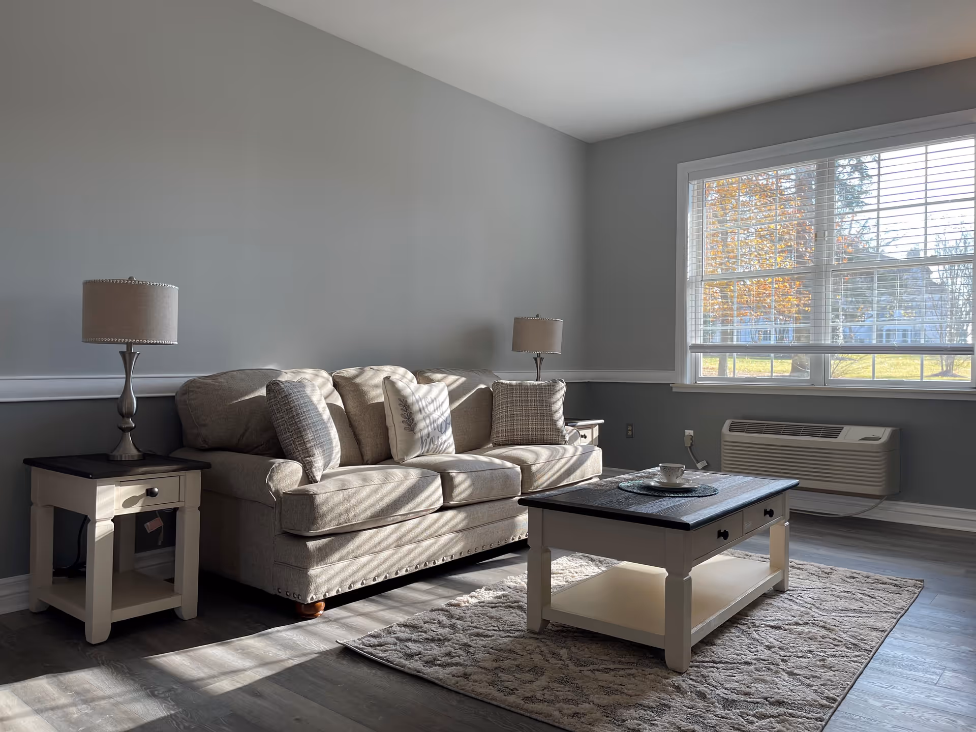 A bright living room with a beige sofa adorned with three decorative pillows, flanked by two matching side tables each holding a lamp. In front of the sofa is a white coffee table with a dark top placed on a patterned rug. A large window with blinds allows natural light to fill the room, and an air conditioning unit is installed below the window.