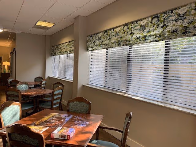 A dining area with several wooden tables and chairs with green upholstery. Large windows with white blinds and floral valances let in natural light. The room has a beige wall color and a ceiling with recessed lighting.