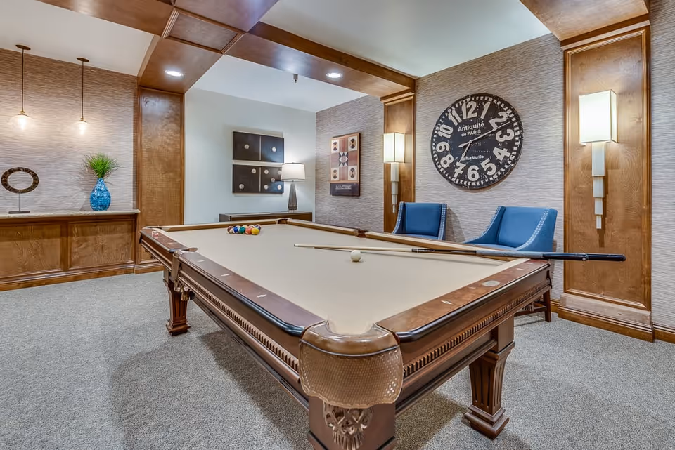 Interior view of a senior living facility game room featuring a pool table with balls and a cue stick on it. The room has carpeted flooring, wooden paneling, two blue chairs, wall-mounted lights, decorative wall art, and a large round clock on the wall.