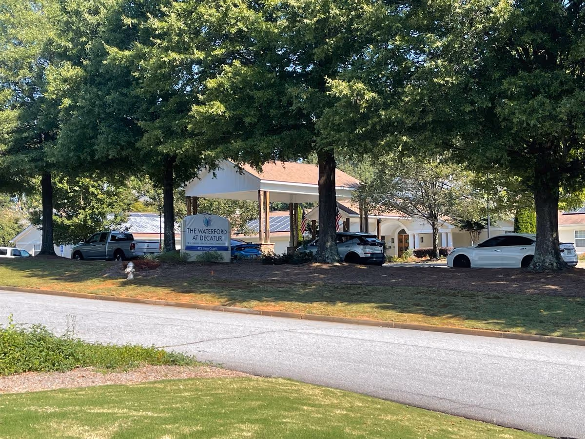 Front entrance of The Waterford at Decatur senior living facility with a driveway, parked cars, large trees, and a sign.