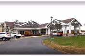 Exterior view of a single-story building with a covered entrance and several parked cars in front, surrounded by a paved driveway and grassy areas under an overcast sky.
