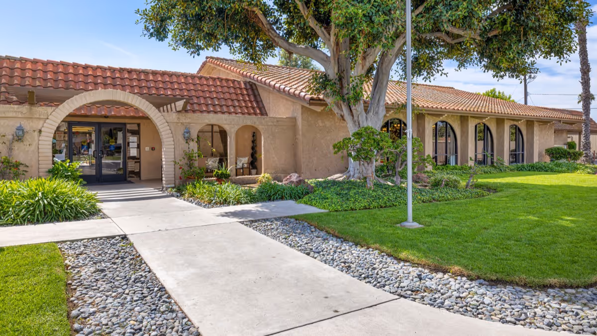 Exterior view of Brookdale Garden Grove facility showing a single-story building with a tiled roof, arched entrance, large windows, well-maintained green lawn, trees, and a concrete walkway leading to the entrance.