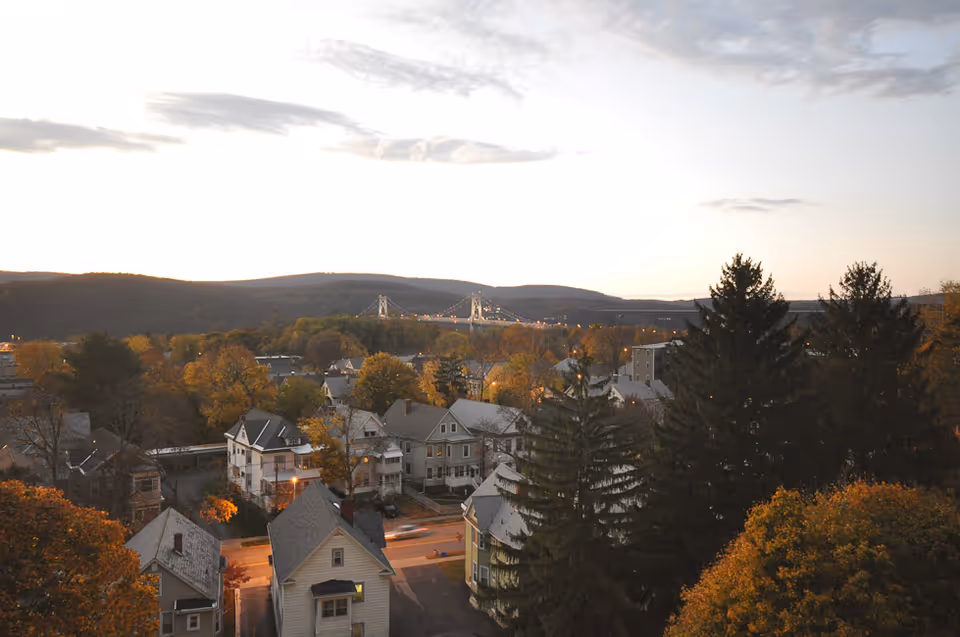 A scenic view of a residential neighborhood with houses and trees in autumn colors, hills in the background, and a suspension bridge visible in the distance under a partly cloudy sky during sunset.