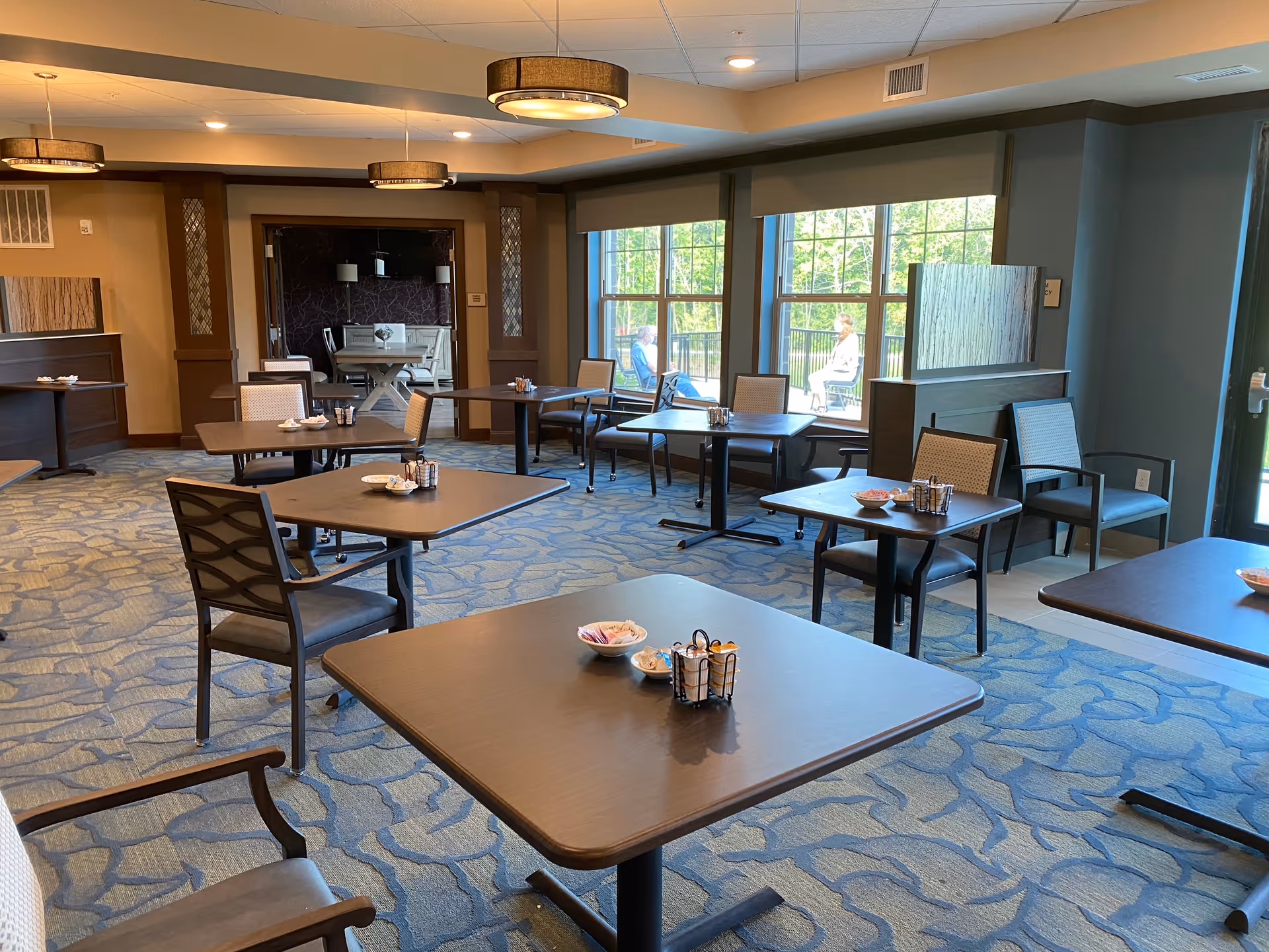 Spacious dining room with multiple square tables and chairs, carpeted floor, and large windows letting in natural light.
