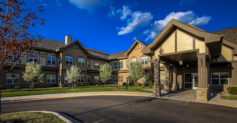 Exterior front view of a two-story senior living facility building under a blue sky with scattered clouds. The building features beige and brown tones with multiple windows and a covered entrance supported by stone pillars. Small trees and landscaping are visible in front of the building along a curved driveway.