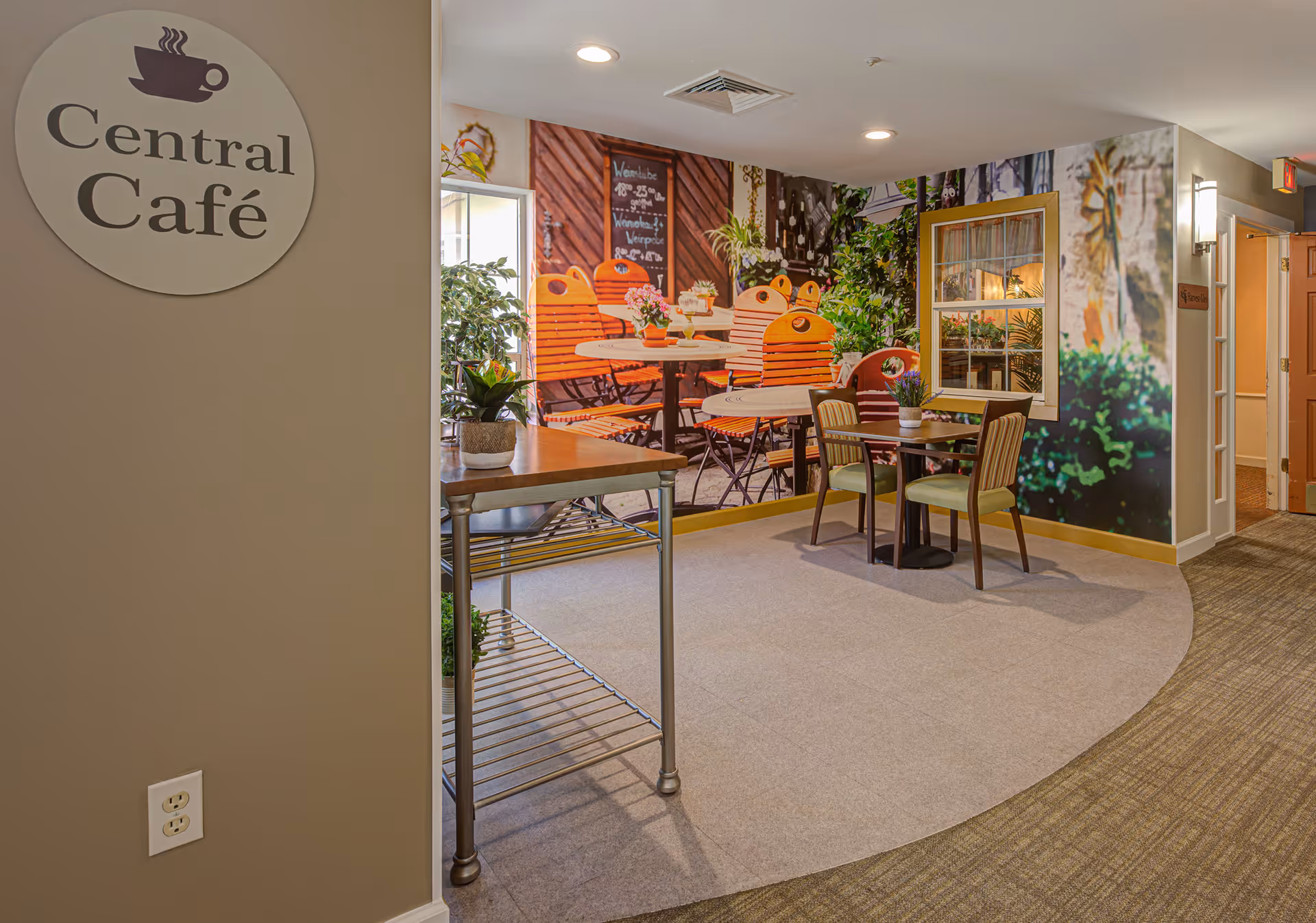 Interior view of the Central Café area in a senior living facility, featuring a small dining table with two chairs, potted plants, and a large wall mural depicting an outdoor café scene with orange chairs and tables. The space has carpeted flooring and soft lighting.