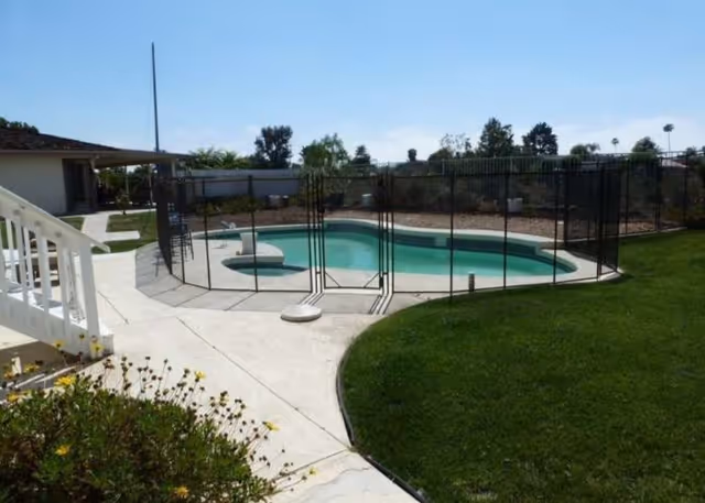 Kidney-shaped outdoor swimming pool with a black safety fence, concrete patio, and surrounding lawn and landscaping.