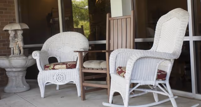 Outdoor patio area with two white wicker chairs with floral cushions, a wooden chair with a cushion, and a decorative stone fountain with a sculpture of two children holding an umbrella.