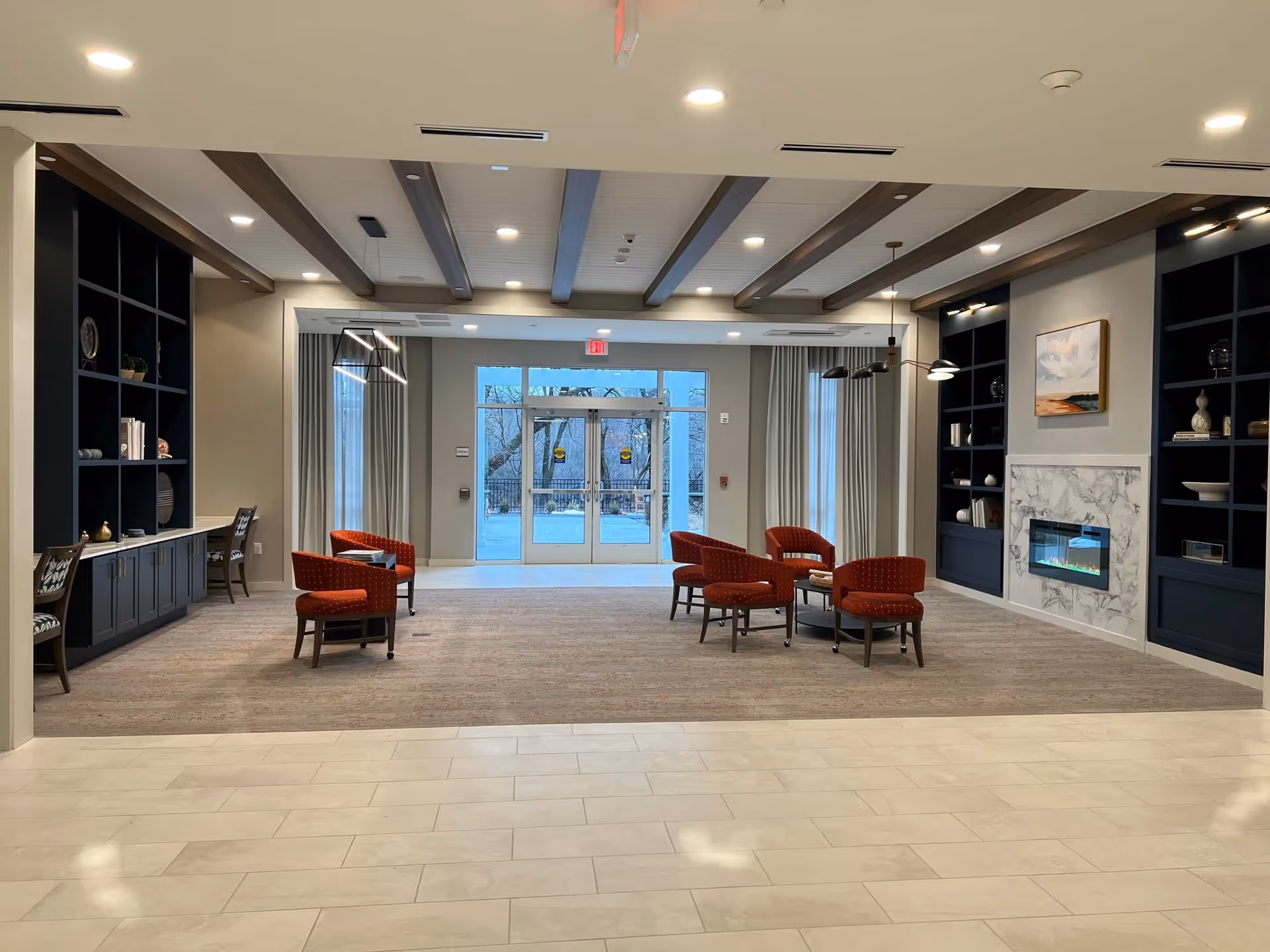 A spacious and modern common area with beige tiled floor in the foreground and carpeted seating area in the background. The seating area features six red upholstered chairs arranged around two small round tables. On the right side, there is a white marble fireplace flanked by built-in dark blue shelves with decorative items and a painting above the fireplace. On the left side, there are built-in desks with chairs and shelves above them. The ceiling has exposed wooden beams and recessed lighting. Large glass doors at the back lead outside, with curtains on either side.