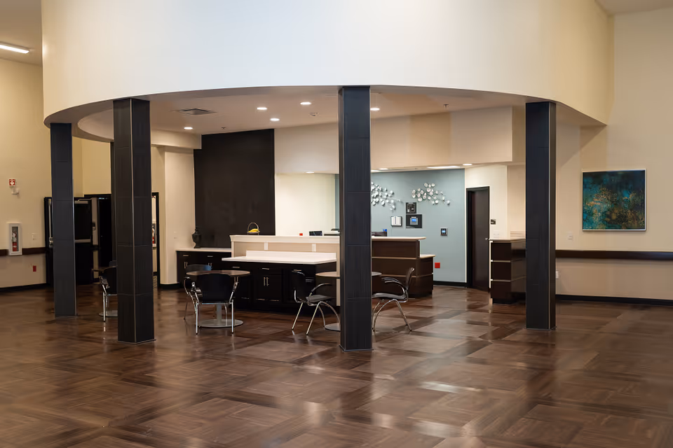 Spacious modern reception lobby with a central curved desk, seating, dark pillars, and wood-patterned flooring.