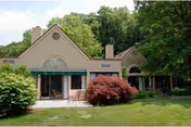 Exterior view of a single-story building with large windows and a patio area, surrounded by green grass, bushes, and trees under a clear sky.