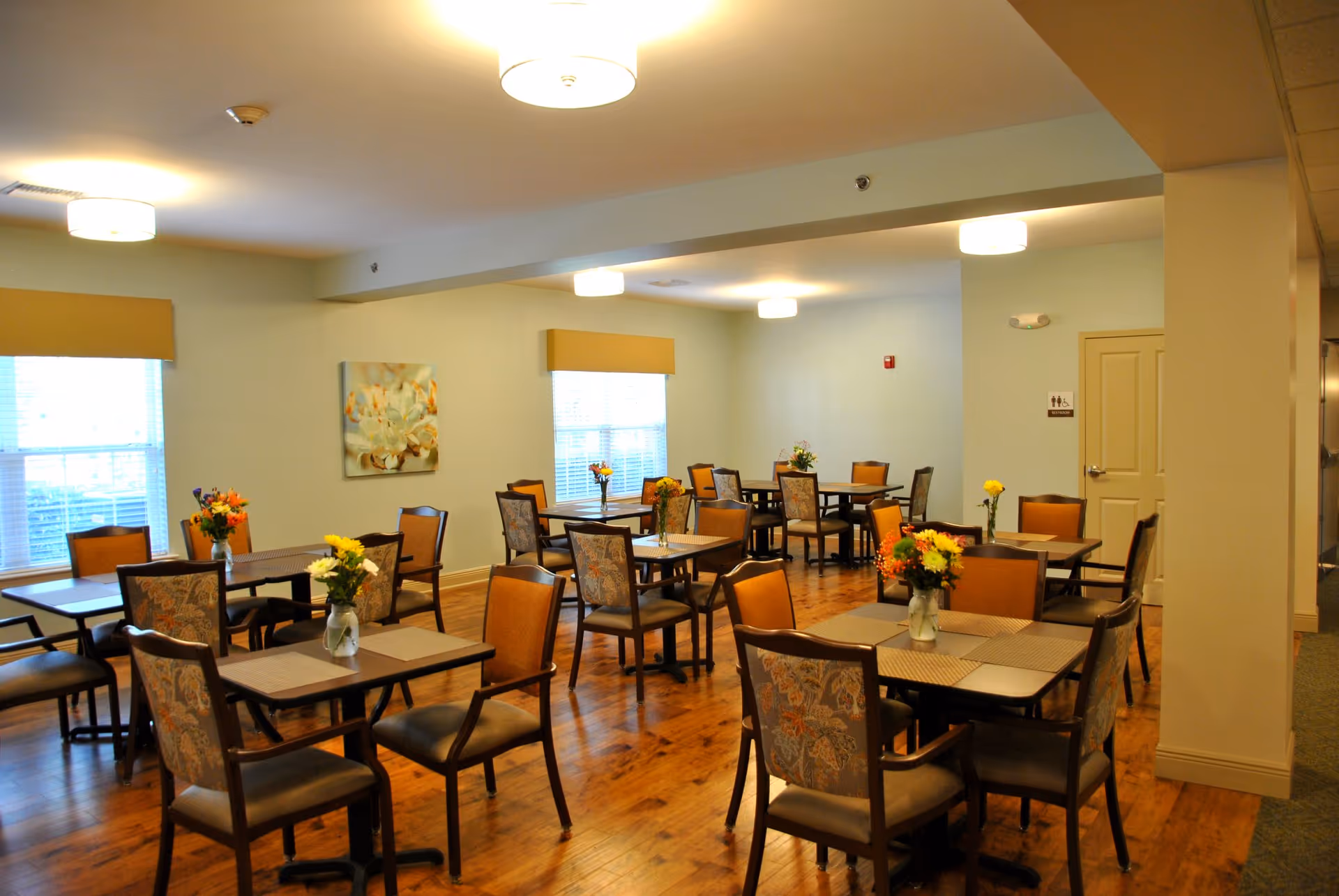 Well-lit dining room with multiple tables and chairs on a hardwood floor, each table decorated with small flower arrangements.