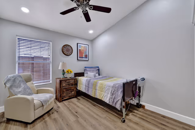 A simple, clean bedroom with a single bed covered in a striped quilt, a wooden nightstand with a lamp and flowers, a beige recliner chair with a white cover, a window with blinds, a wall clock, and a framed abstract artwork. The room has light-colored walls, wood flooring, and a ceiling fan with lights.