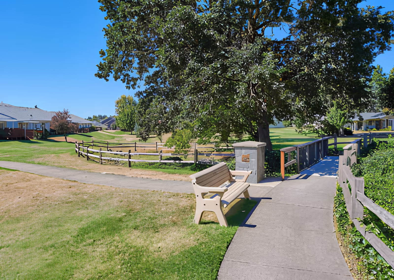 Outdoor area at Elliott Residence featuring a paved walkway with a wooden bench, a small bridge with wooden railings, large trees providing shade, green grass, and residential buildings in the background under a clear blue sky.